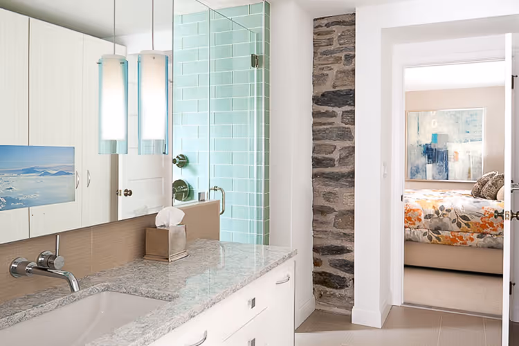Modern bathroom with granite countertop, white cabinets, glass shower with mint green tiles, and doorway showing bedroom with floral bedspread.