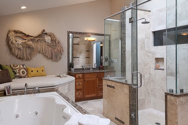 Modern bathroom with a glass-enclosed shower, a large bathtub with towels, wooden vanity with mirror, and cushioned bench with decorative pillows and wall hanging.