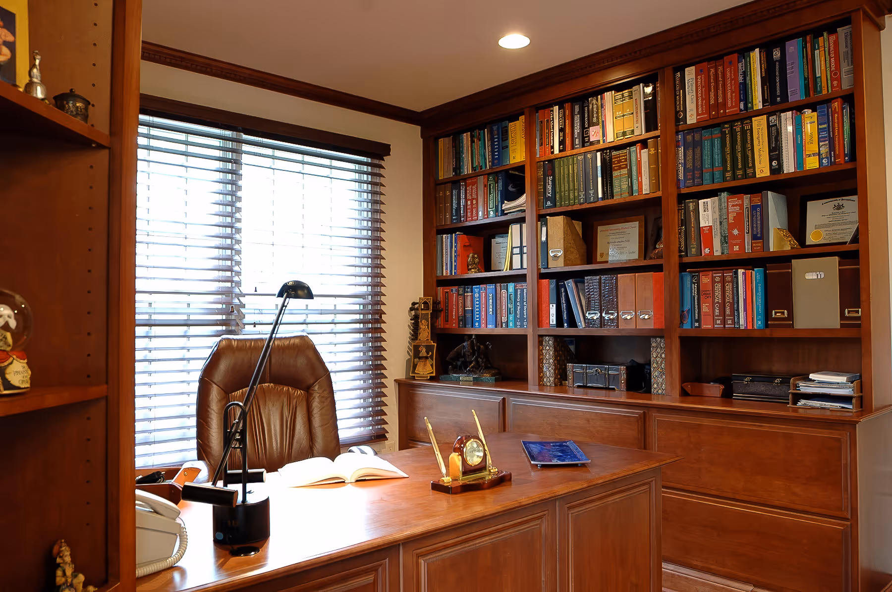 Wood-paneled home office with a large desk, leather chair, desk lamp, open book, and a bookshelf filled with books and documents.
