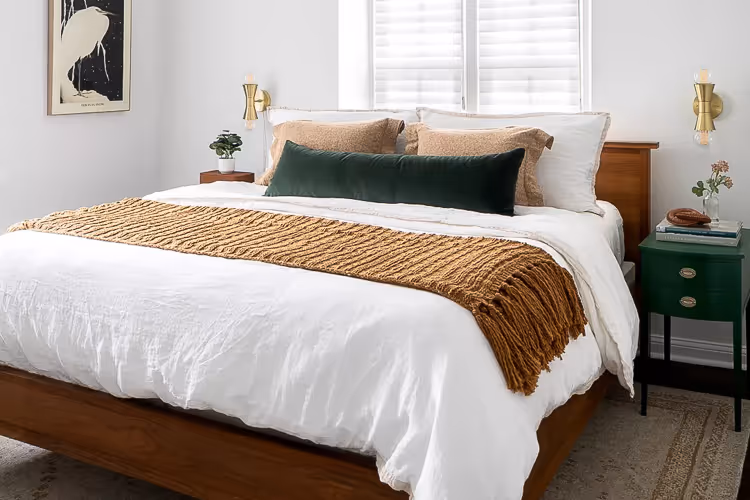Modern bedroom with a wooden bed frame, white bedding, textured brown throw, and green and beige pillows, flanked by green nightstands and wall sconces.
