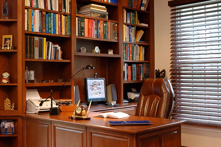 Wooden office desk with a leather chair, open book, and desktop computer against a background of filled wooden bookshelves and window blinds.