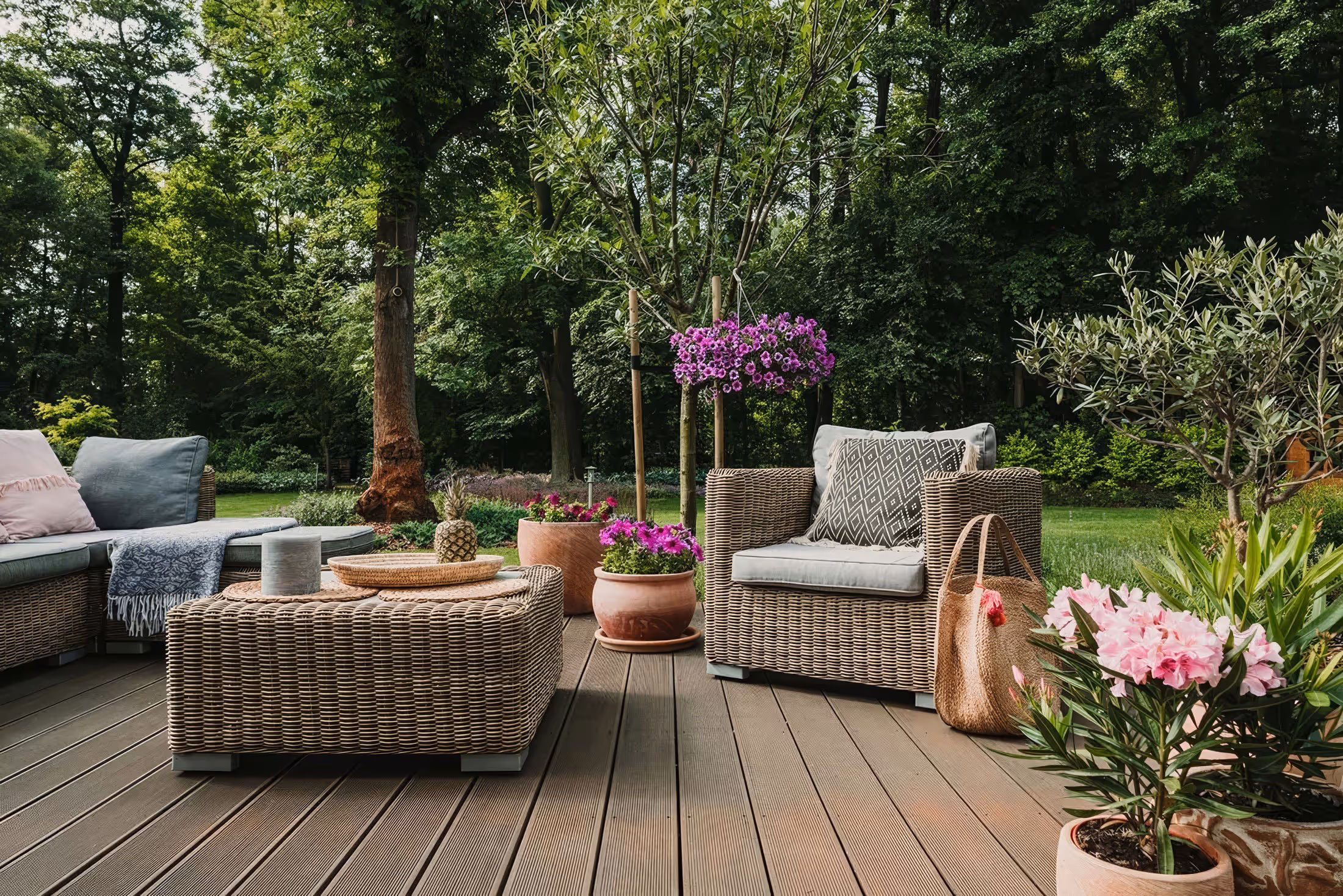 Outdoor patio with wicker furniture, gray cushions, a coffee table with decor, and various potted plants and flowers on a wooden deck surrounded by trees.