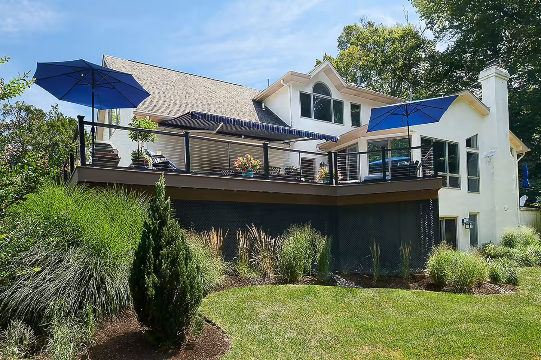 White two-story house with elevated wooden deck featuring black railing, blue umbrellas, and outdoor furniture surrounded by green lawn and shrubs.