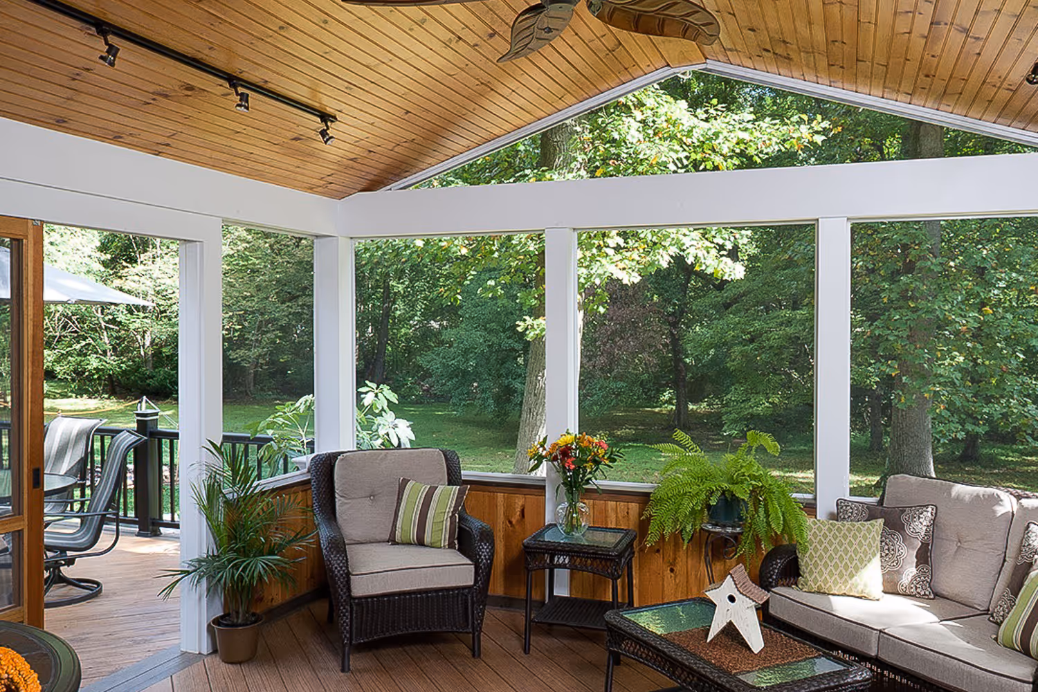 Cozy screened porch with wicker armchair, sofa, glass-top tables, potted plants, and a view of green trees outside.