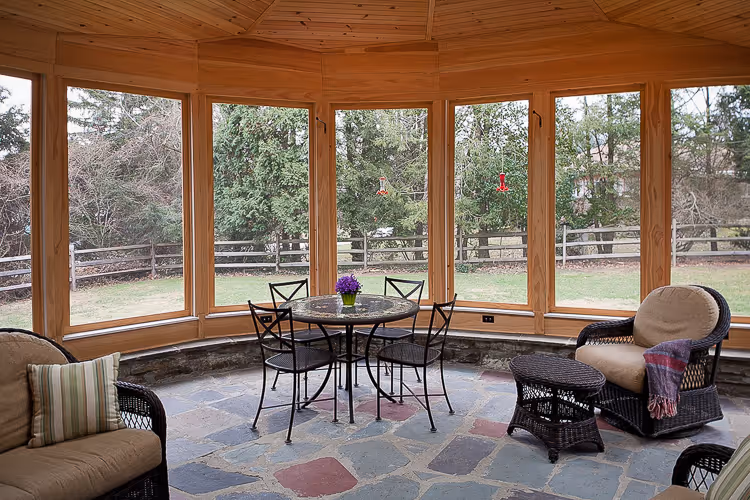 Wooden sunroom with large windows overlooking a backyard, furnished with a round metal table, four chairs, and cushioned wicker armchairs.