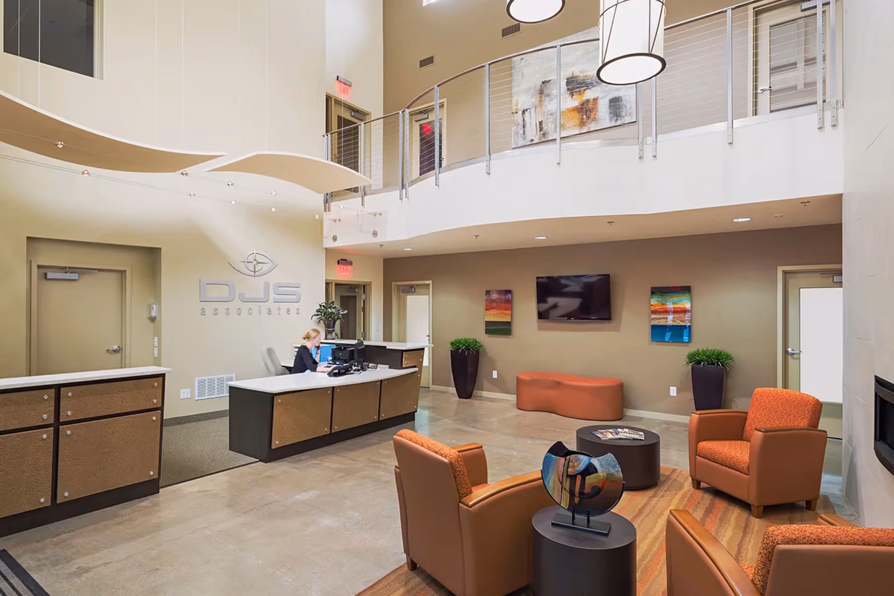 Modern office reception area with a receptionist at desk, orange chairs, abstract artwork, and a wall-mounted TV.