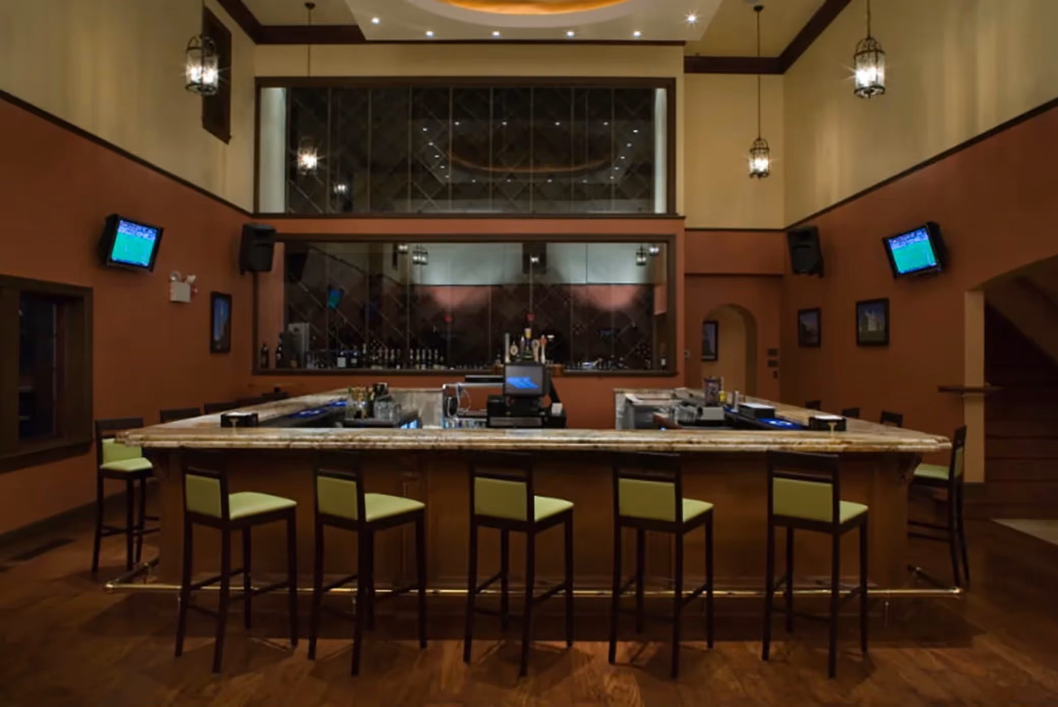 Empty U-shaped bar with green cushioned stools in a warmly lit room featuring televisions on the walls and a mirrored back wall with bottles.