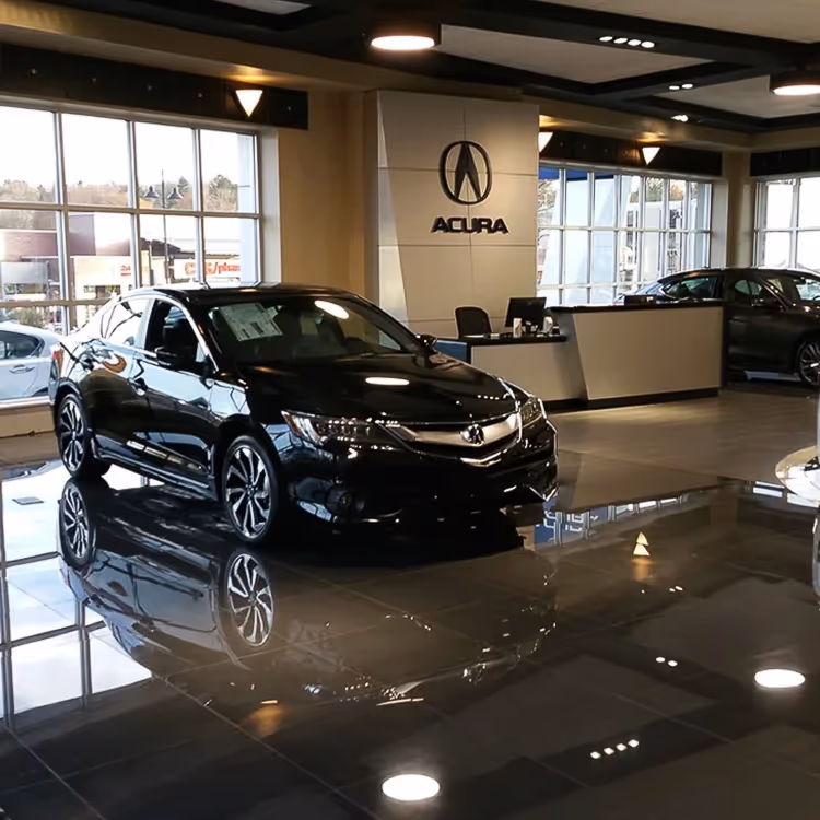 Black Acura sedan displayed inside an Acura dealership showroom with reflective tiled floor and large windows.