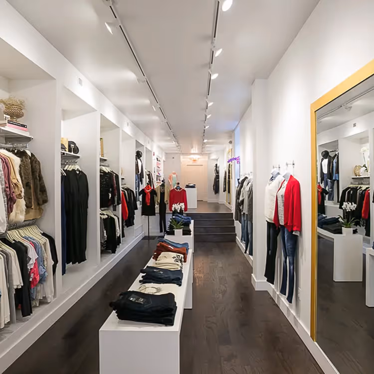 Modern clothing boutique interior with apparel displayed on wall hangers, mannequins, and folded on center tables under bright ceiling lights.