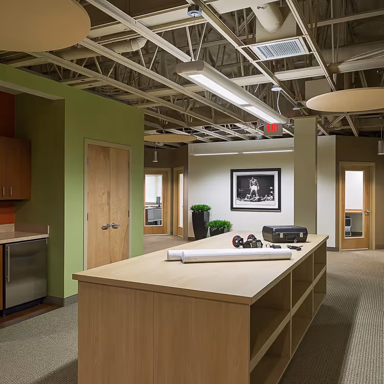 Modern office space with a long wooden table holding rolled blueprints, a toolbox, dumbbells, and a calculator, with green walls and exposed ceiling beams.