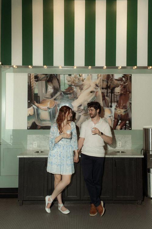 A couple holding hands and smiling at each other while holding coffee cups, standing under green and white striped awning in front of a carousel horse photo.