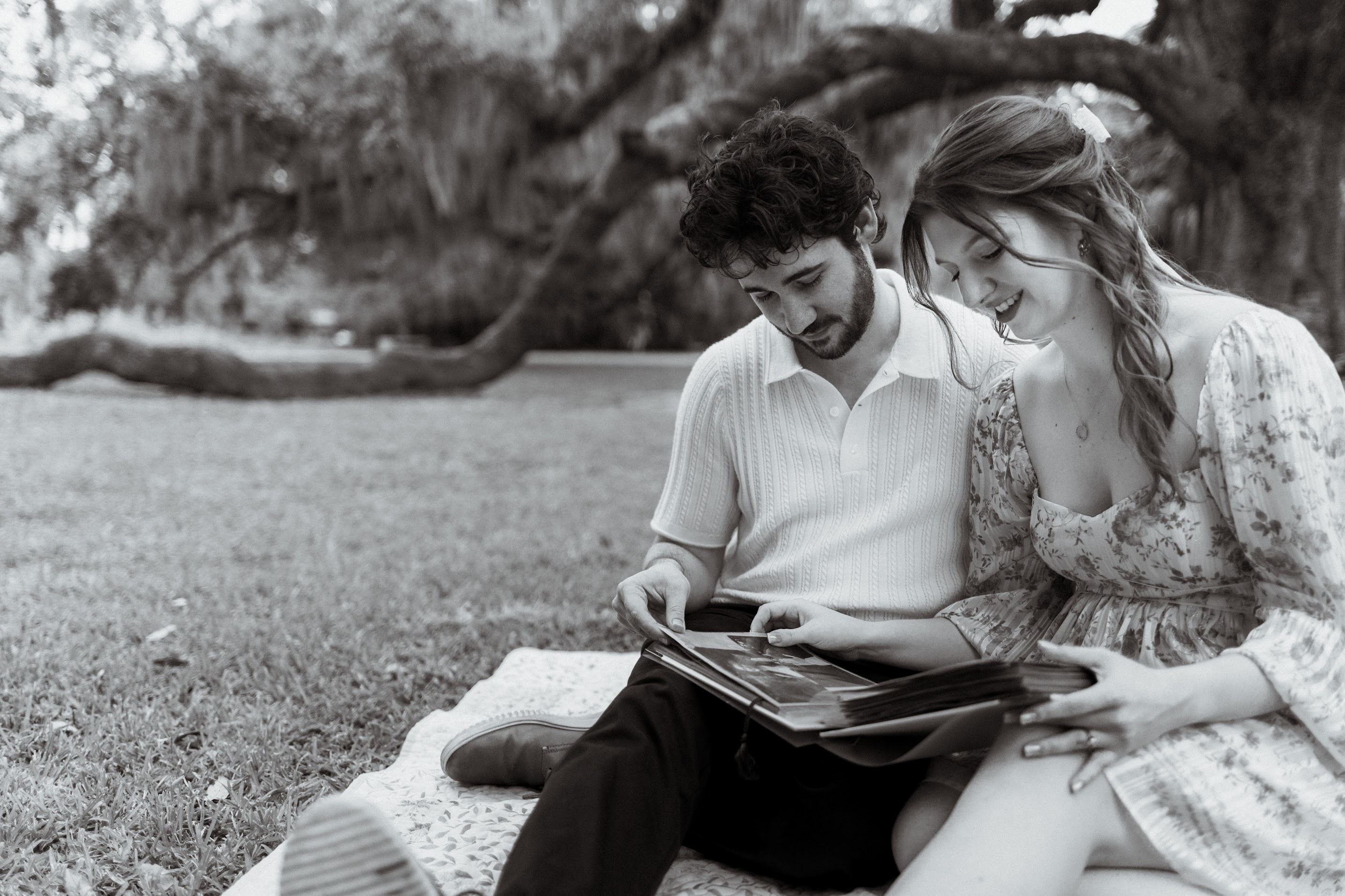 Young couple sitting on a blanket in a grassy park, looking through a photo album together.