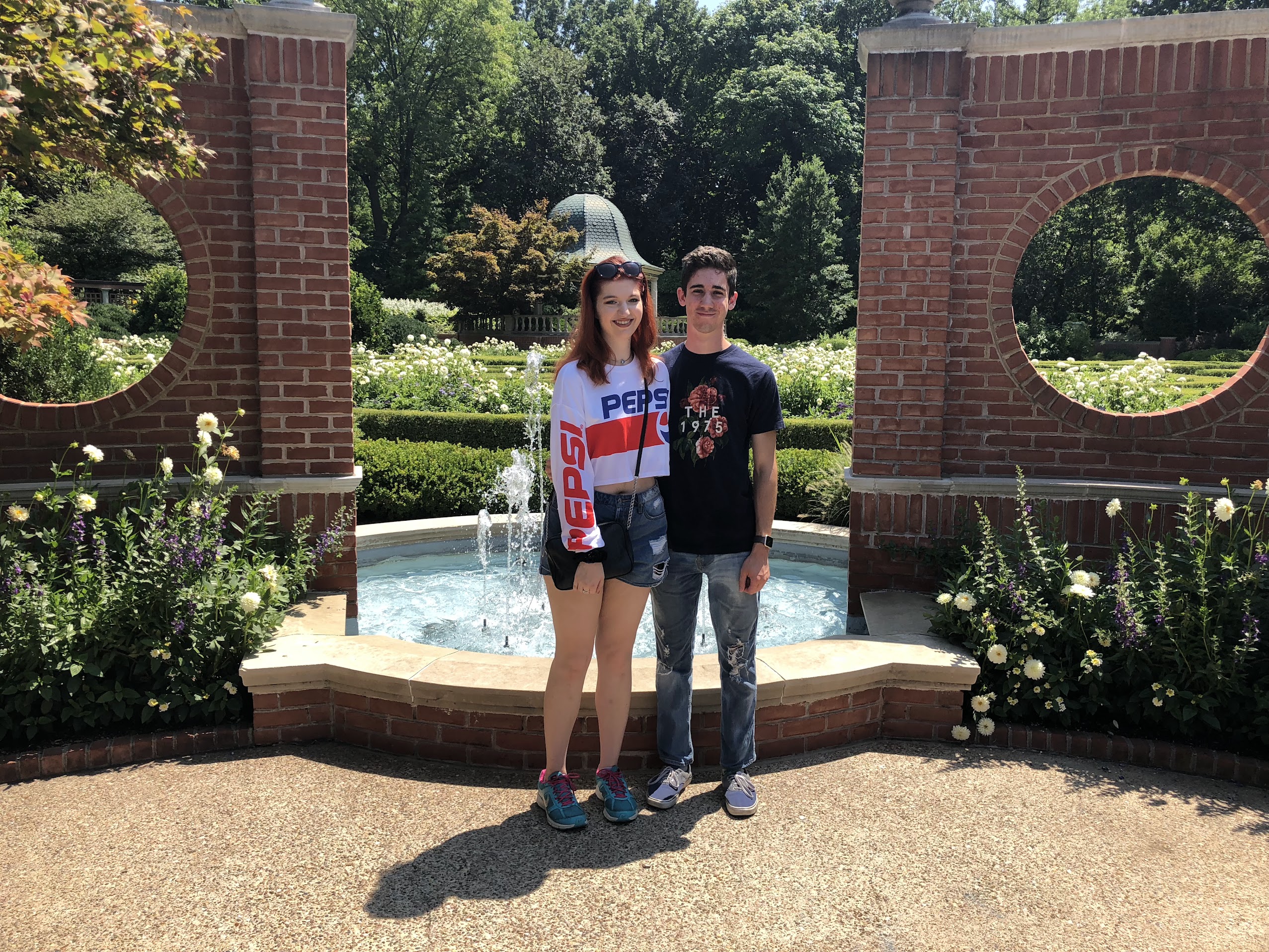 Young woman in a Pepsi sweatshirt and shorts standing next to a young man in a dark t-shirt and jeans in front of a circular garden fountain and brick wall with round openings.