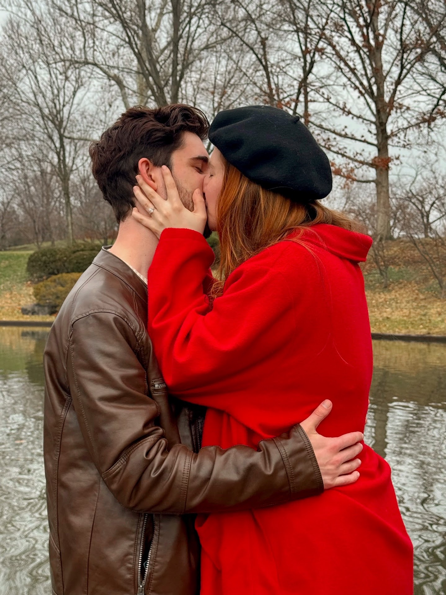 Couple kissing outdoors near a pond with bare trees in the background; woman wearing red coat and black beret, man wearing brown leather jacket.