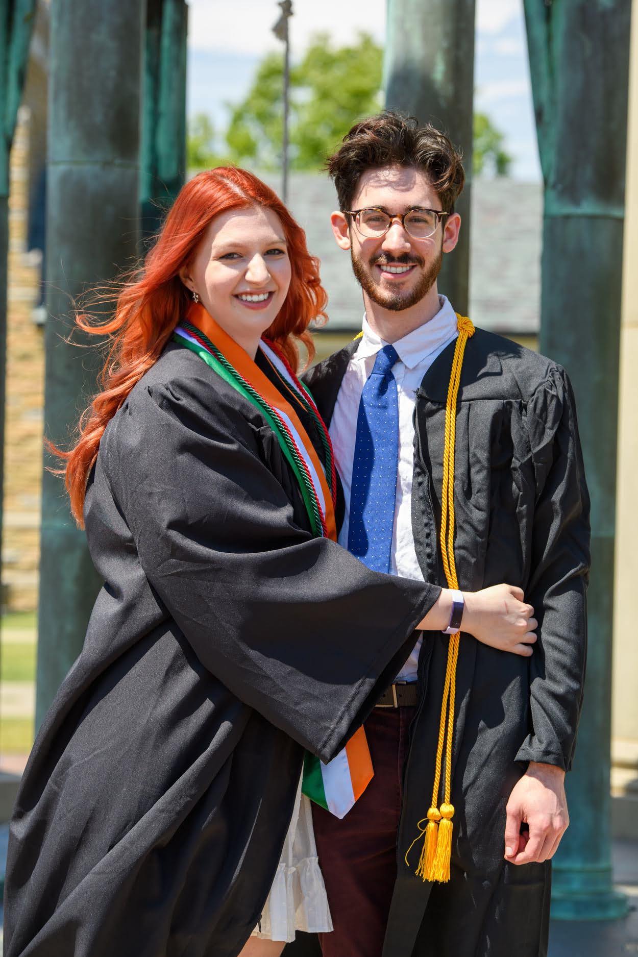 Smiling young man and woman wearing black graduation gowns hugging outdoors near stone columns.