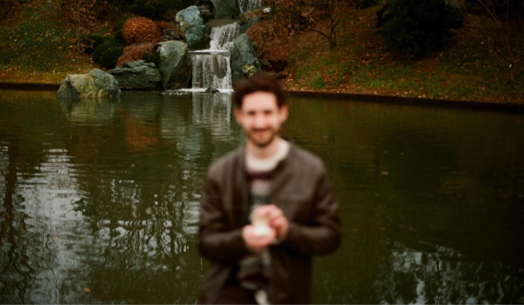 Blurred image of a man holding a small object in front of a pond with a waterfall and rocks.