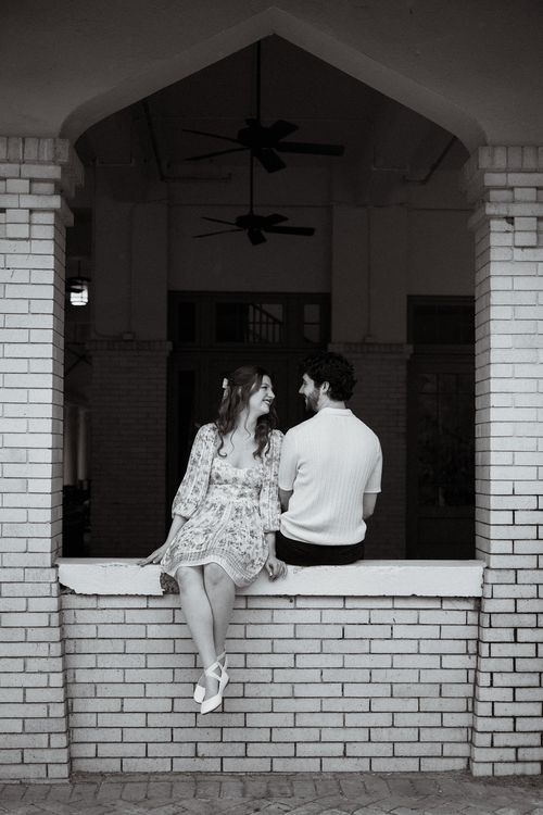Black-and-white photo of a woman in a floral dress and ballet flats sitting on a brick ledge smiling at a man in a short-sleeve sweater who sits beside her with his back turned.