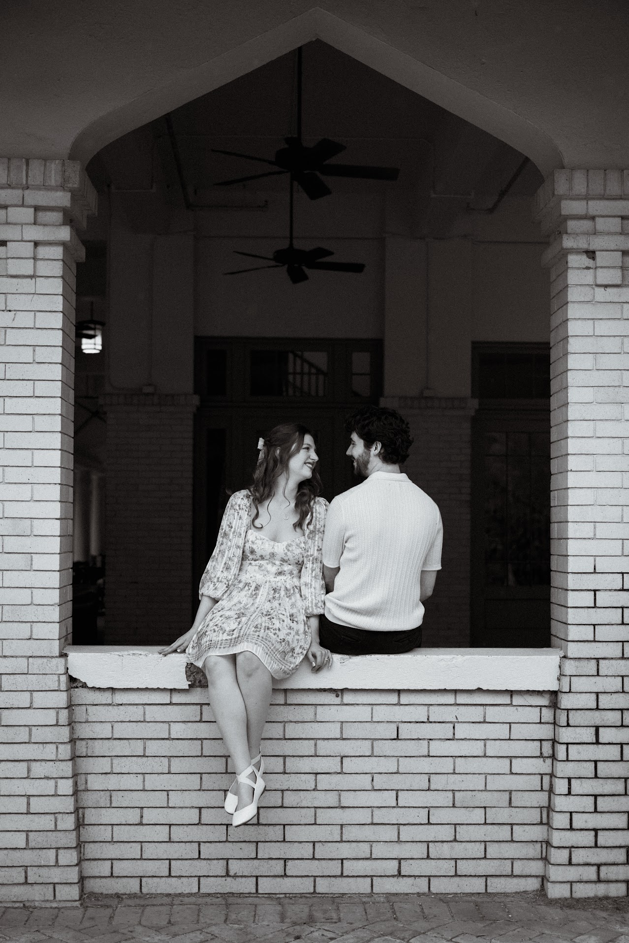 Black-and-white photo of a woman in a floral dress and ballet flats sitting on a brick ledge smiling at a man in a short-sleeve sweater who sits beside her with his back turned.