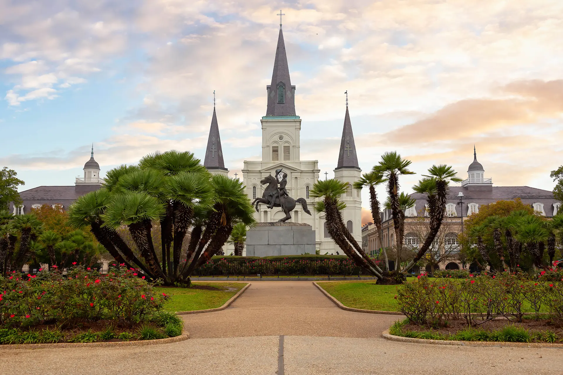 Equestrian statue in front of St. Louis Cathedral with palm trees and gardens at dusk in a city square.