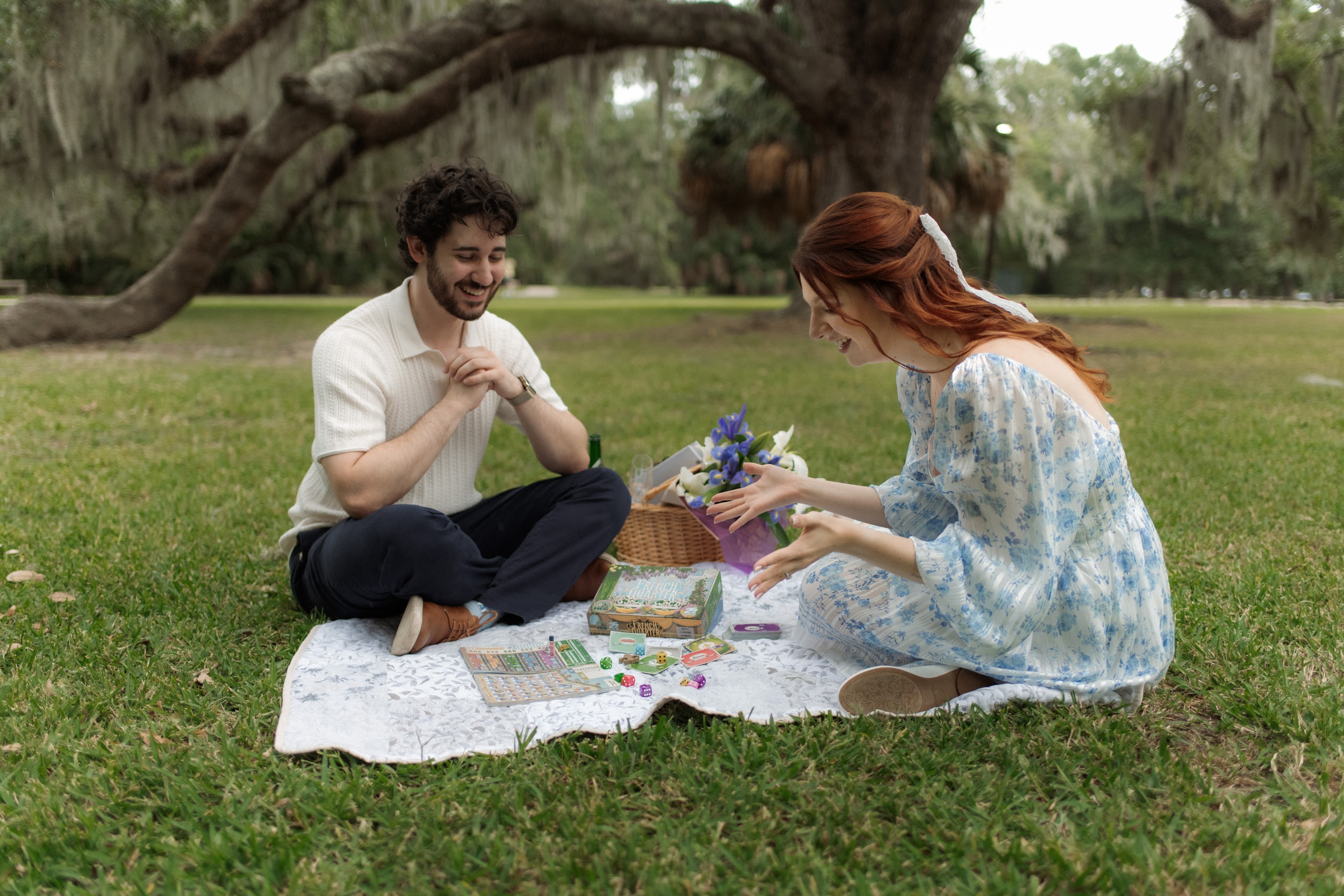 A man and a woman sitting on a blanket having a picnic and playing a board game on the grass under large trees.