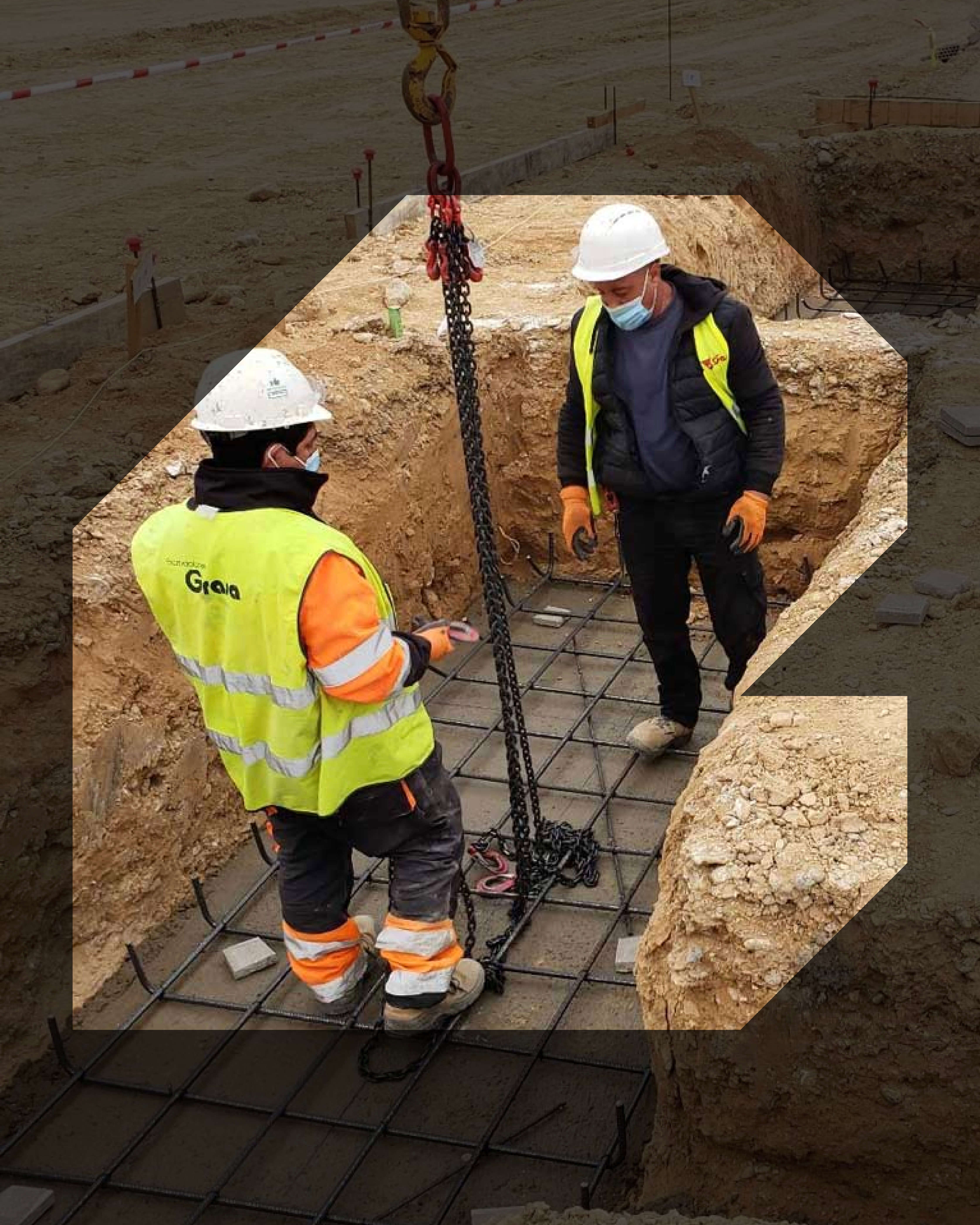 Two construction workers wearing helmets and high-visibility vests stand in a trench handling lifting chains over a steel rebar foundation grid.