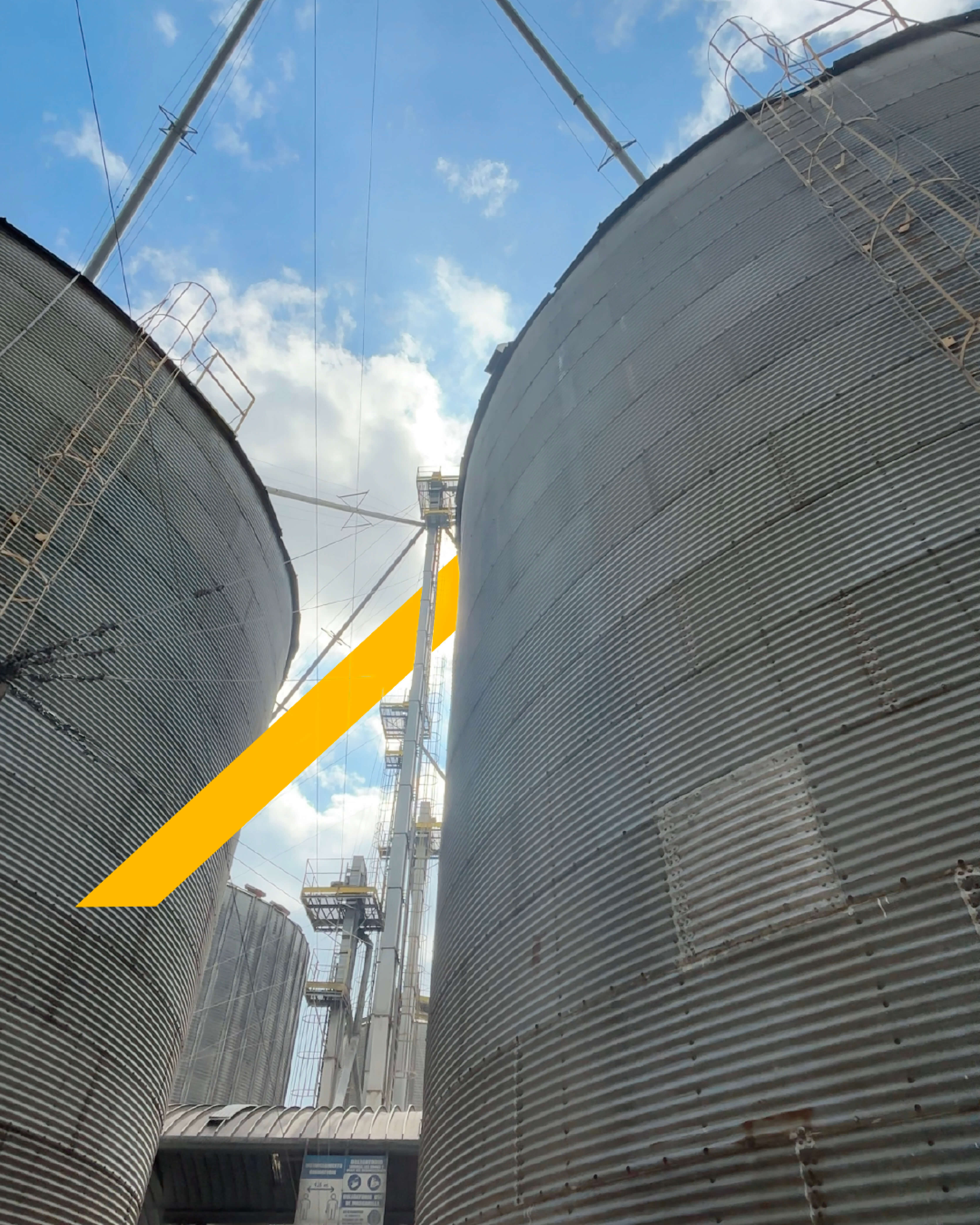 Close-up view looking up between large corrugated metal silos with ladders and metal framework under a partly cloudy blue sky.