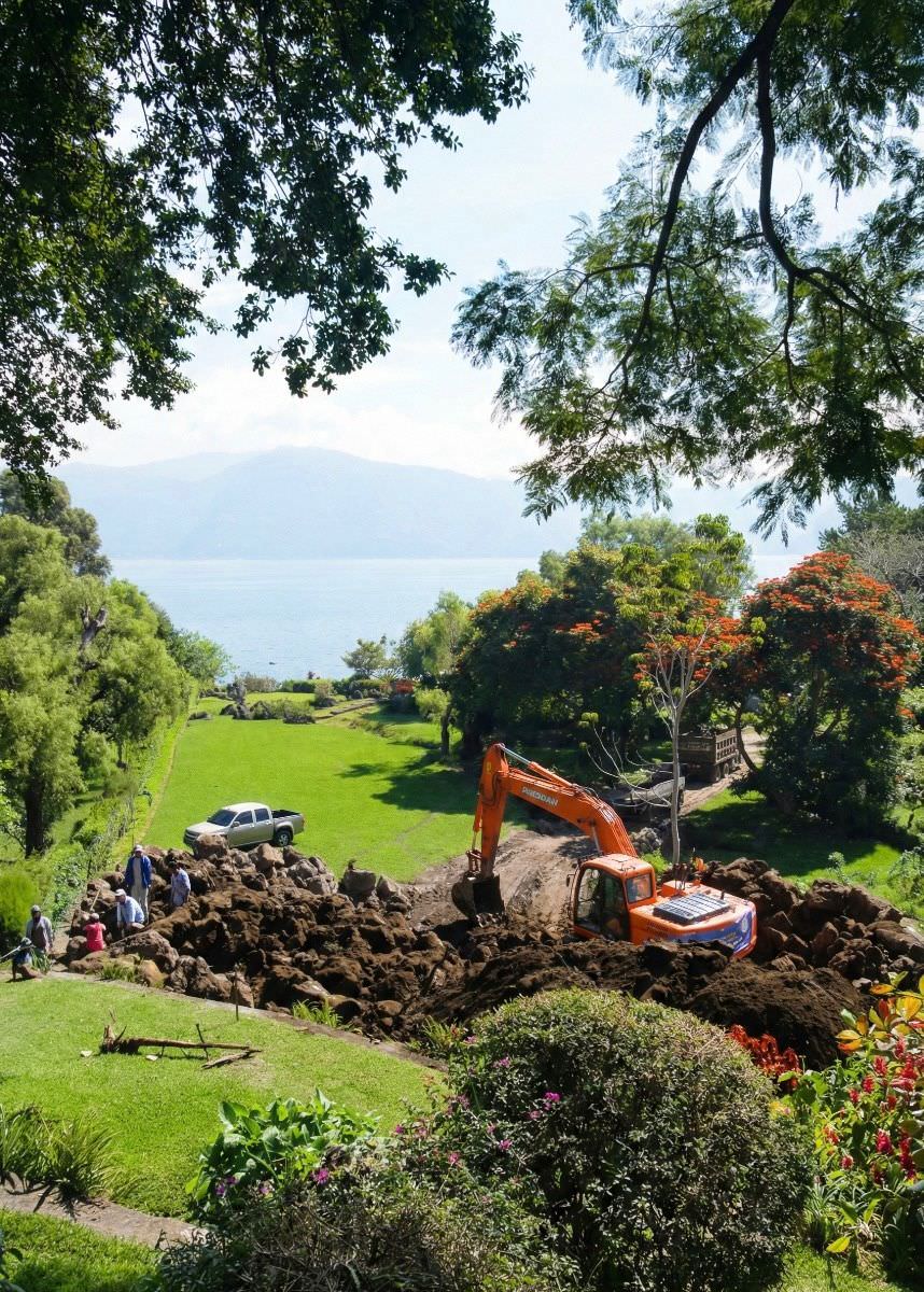 Orange excavator digging soil on green grassy hill with trees and a lake in the background under a partly cloudy sky.