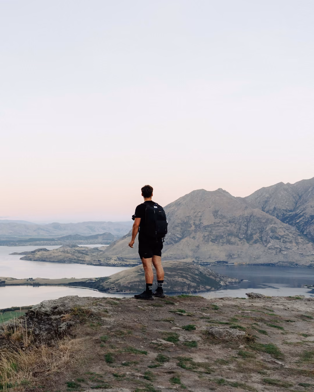 Person standing on a large rock overlooking a lake surrounded by forested hills at sunset.
