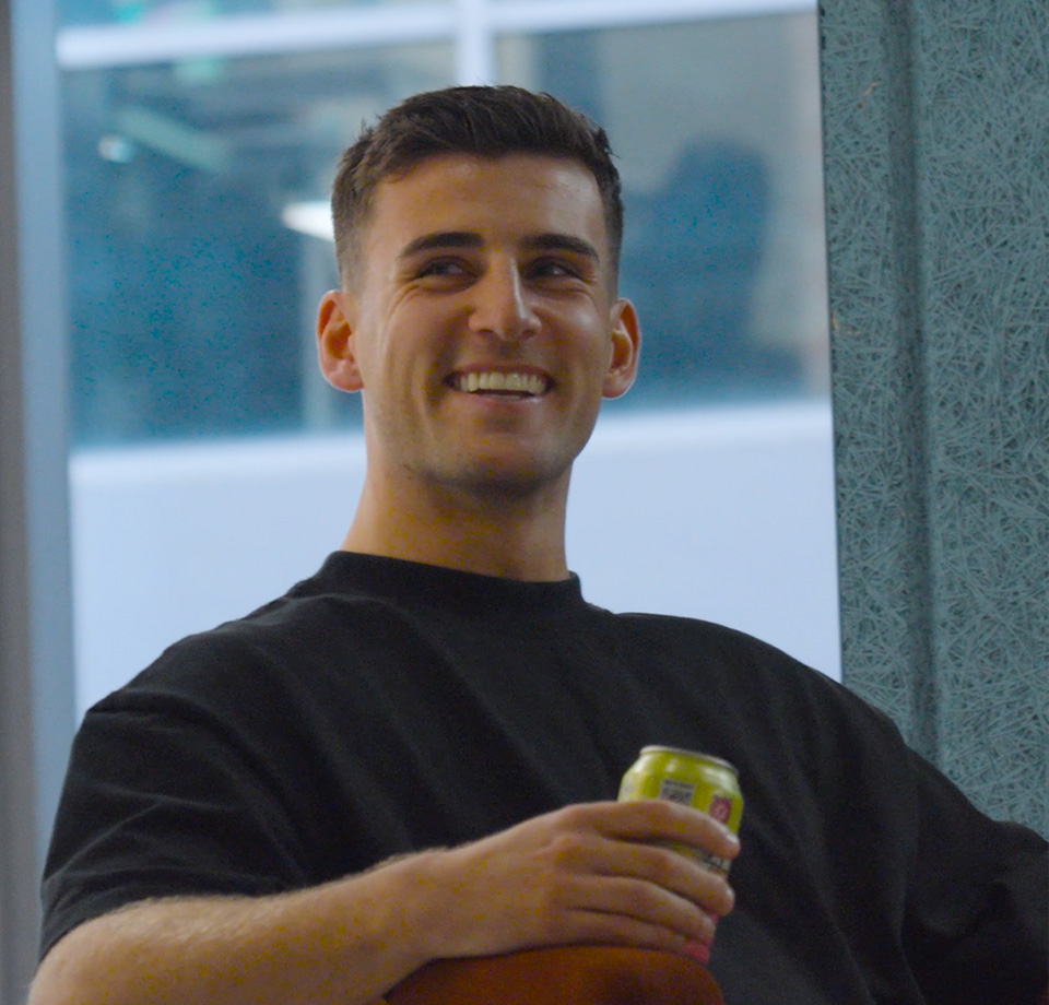 Smiling young man with short dark hair holding a beverage can indoors near a window.