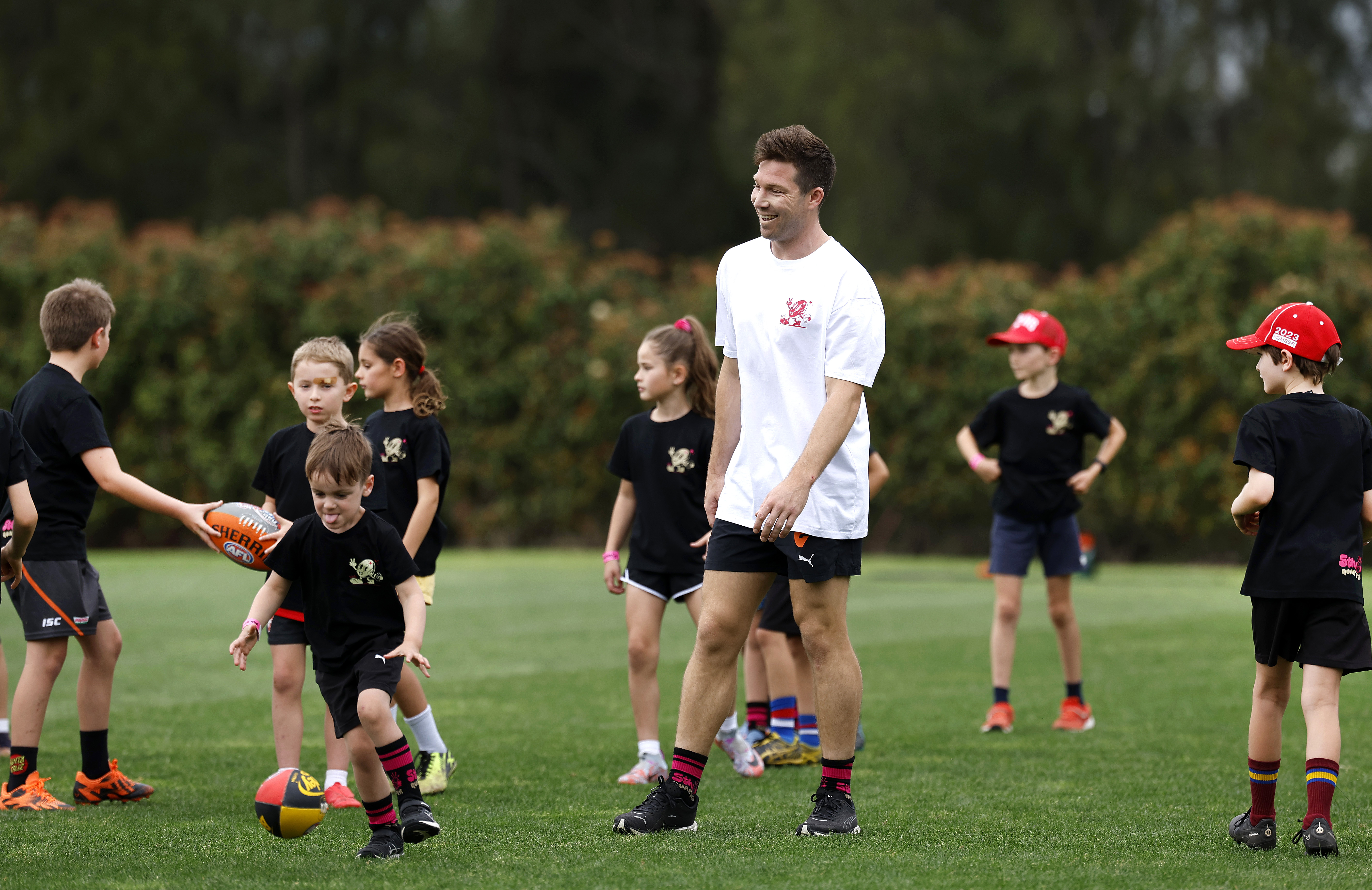 Coach in white shirt smiling with children in black sportswear playing with footballs on grass field.