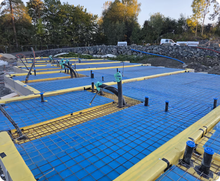 Construction site with a concrete foundation framework featuring blue waterproofing sheets, yellow insulation, and exposed rebar and pipes.
