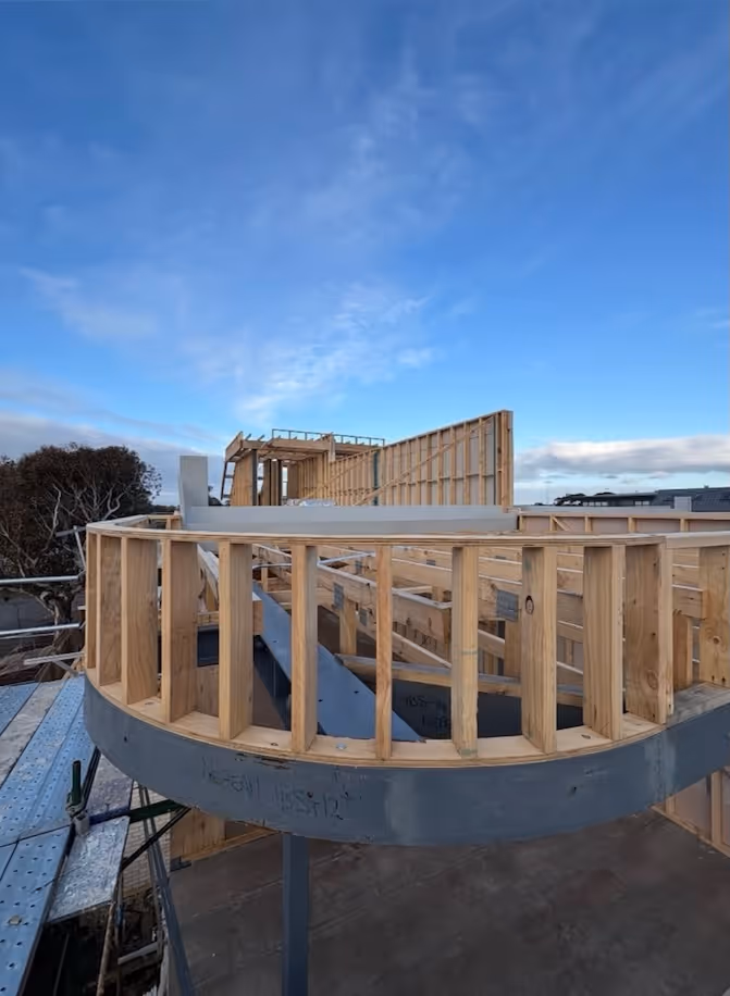 Wooden framing of a modern curved balcony under construction against a blue sky.