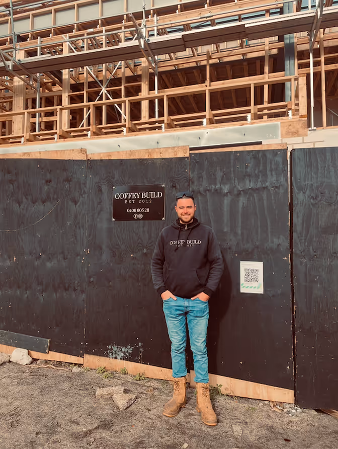 Man wearing a black Coffey Build hoodie and tan boots, standing in front of a black wooden construction site fence with a building framework behind.
