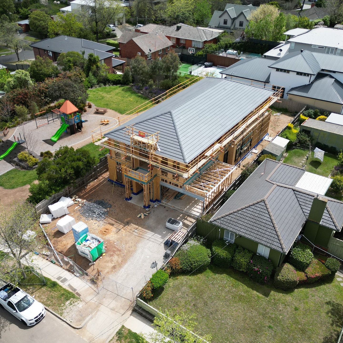 Aerial view of a large house under construction with scaffolding around it, next to completed houses and a playground with green slides.