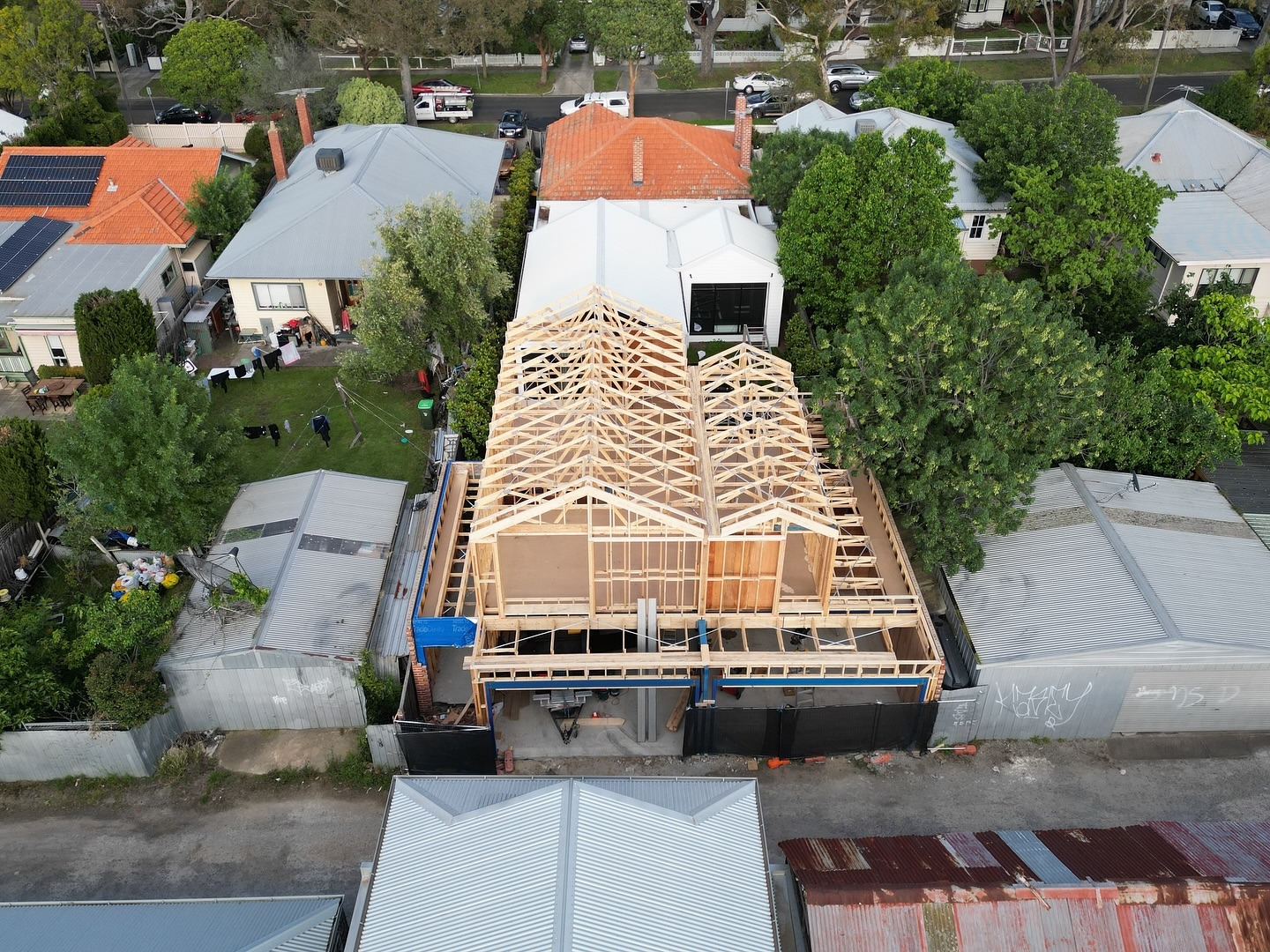 Aerial view of a house under construction with wooden roof framing surrounded by completed homes and green trees.