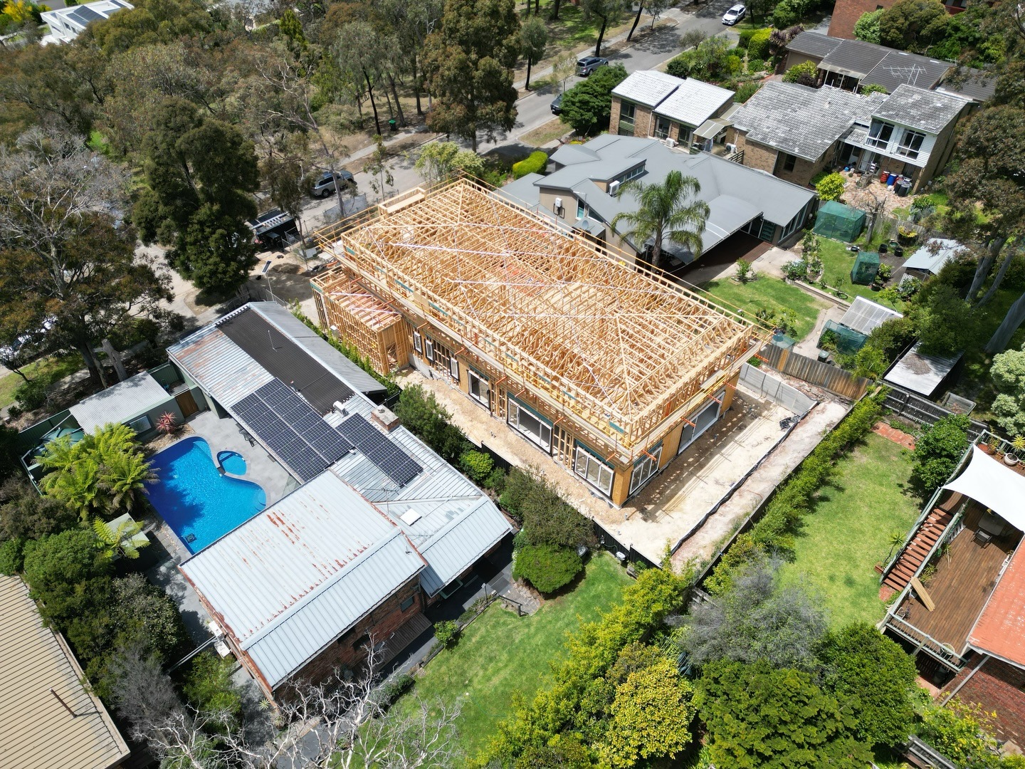 Aerial view of a residential neighborhood featuring a large house under wooden frame construction surrounded by completed houses with gardens and a swimming pool.