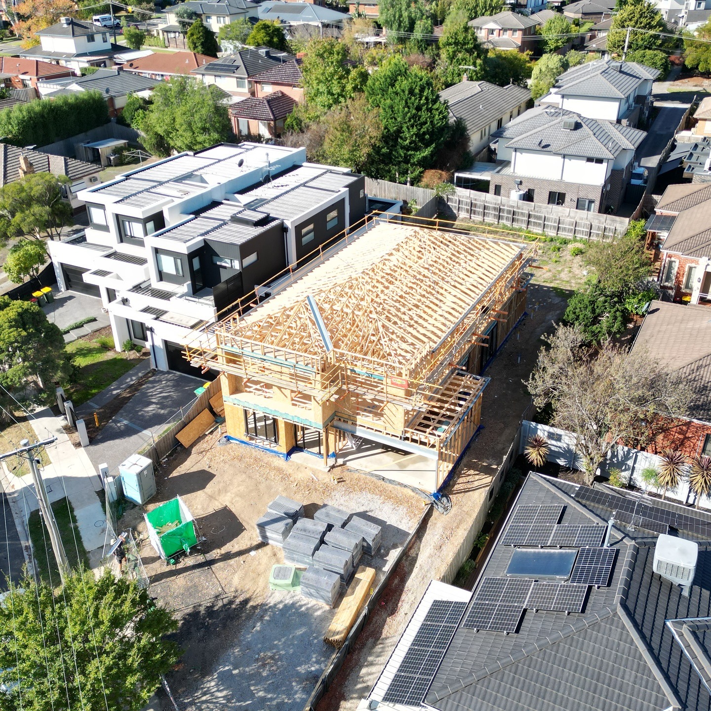 Aerial view of a wooden house under construction in a residential neighborhood with finished houses and solar panels nearby.