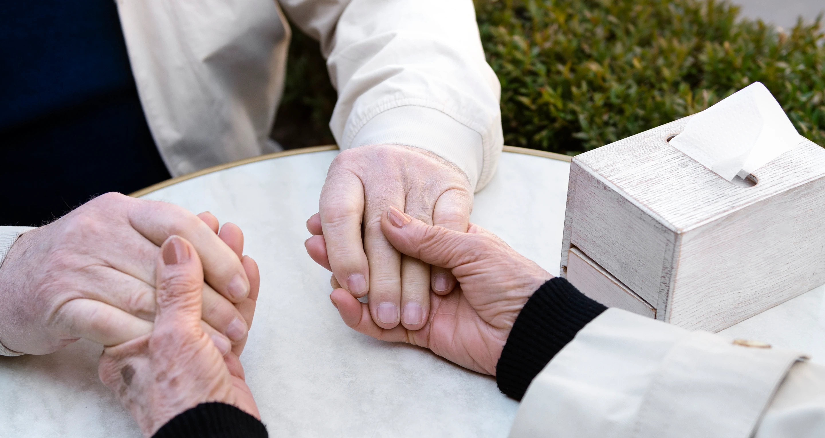 An elderly person holding hands with another person across a round white table with a wooden tissue box nearby.