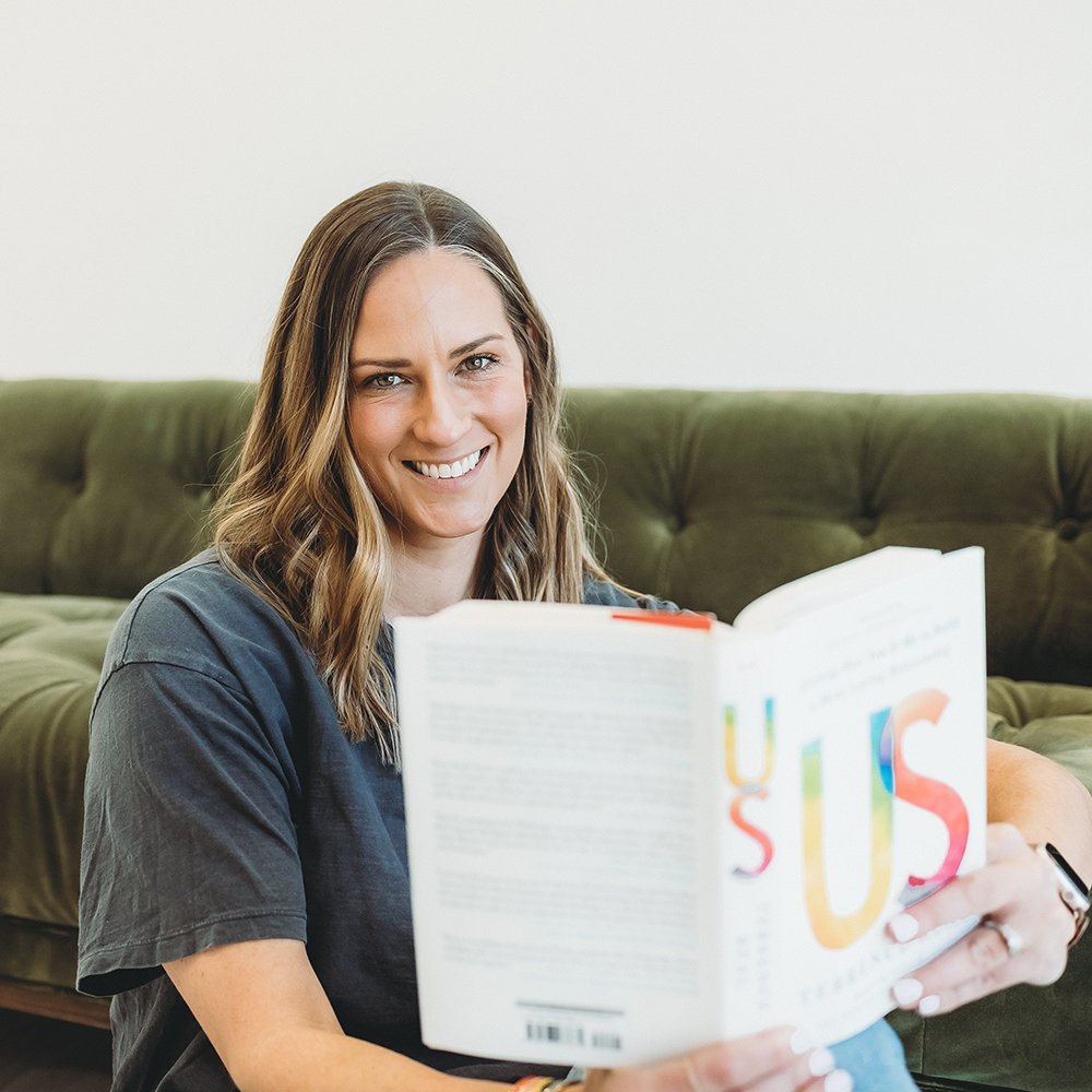 Smiling woman with brown hair sitting on a green couch, holding and reading a book titled 'US'.