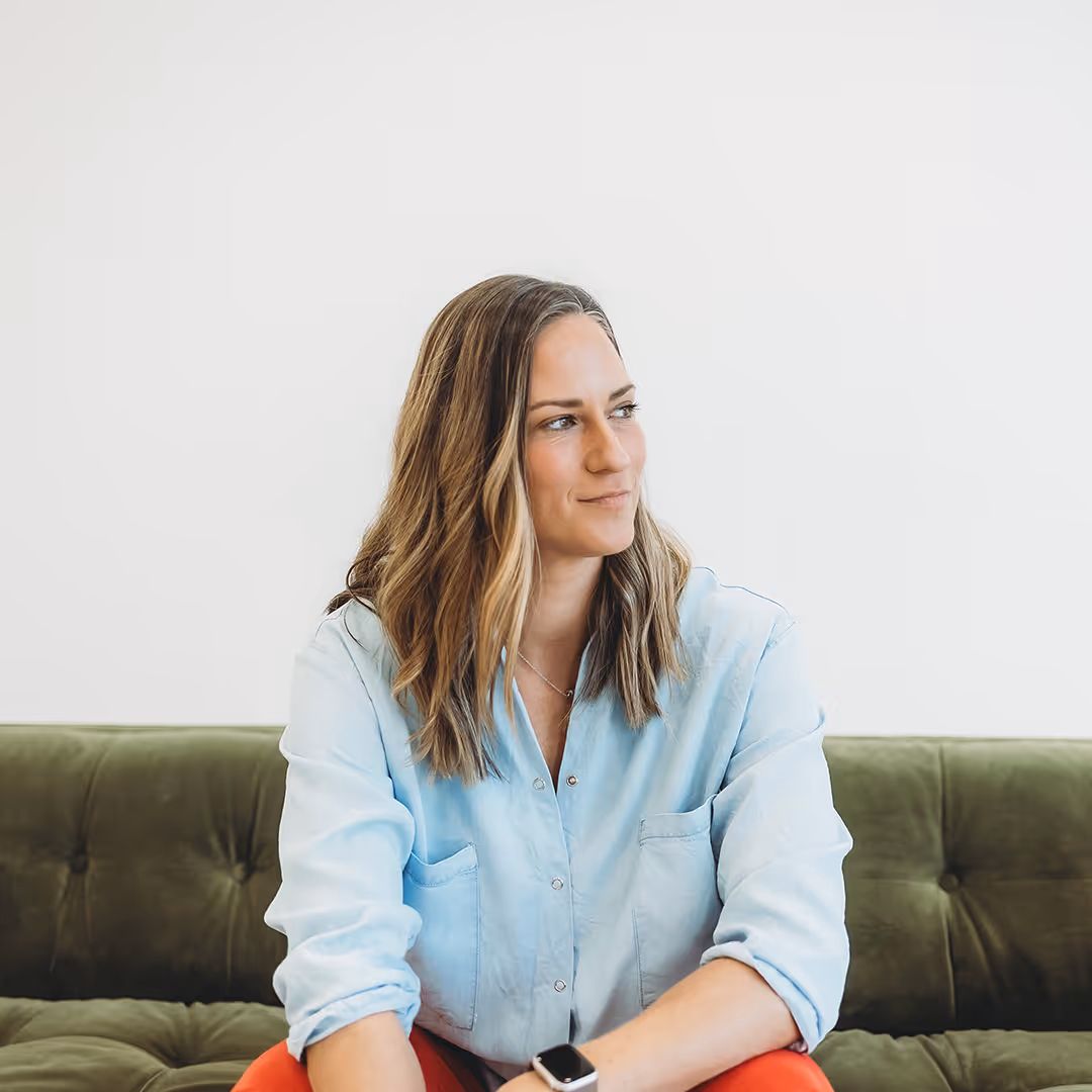 Woman with wavy light brown hair, wearing a light blue shirt and red pants, sitting on a green couch and looking to the side.