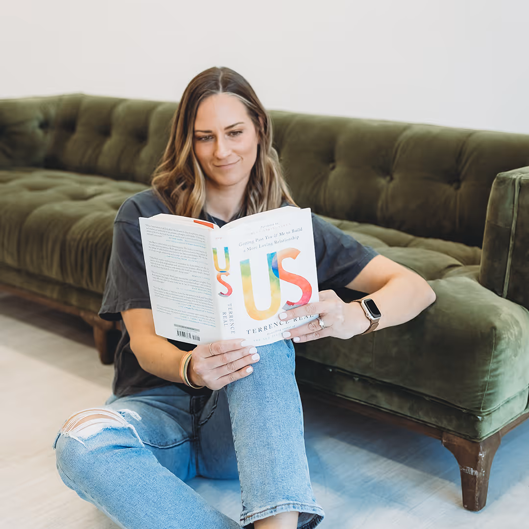 Woman with wavy hair sitting on the floor by a green couch, reading the book 'Us' by Terrence Real.