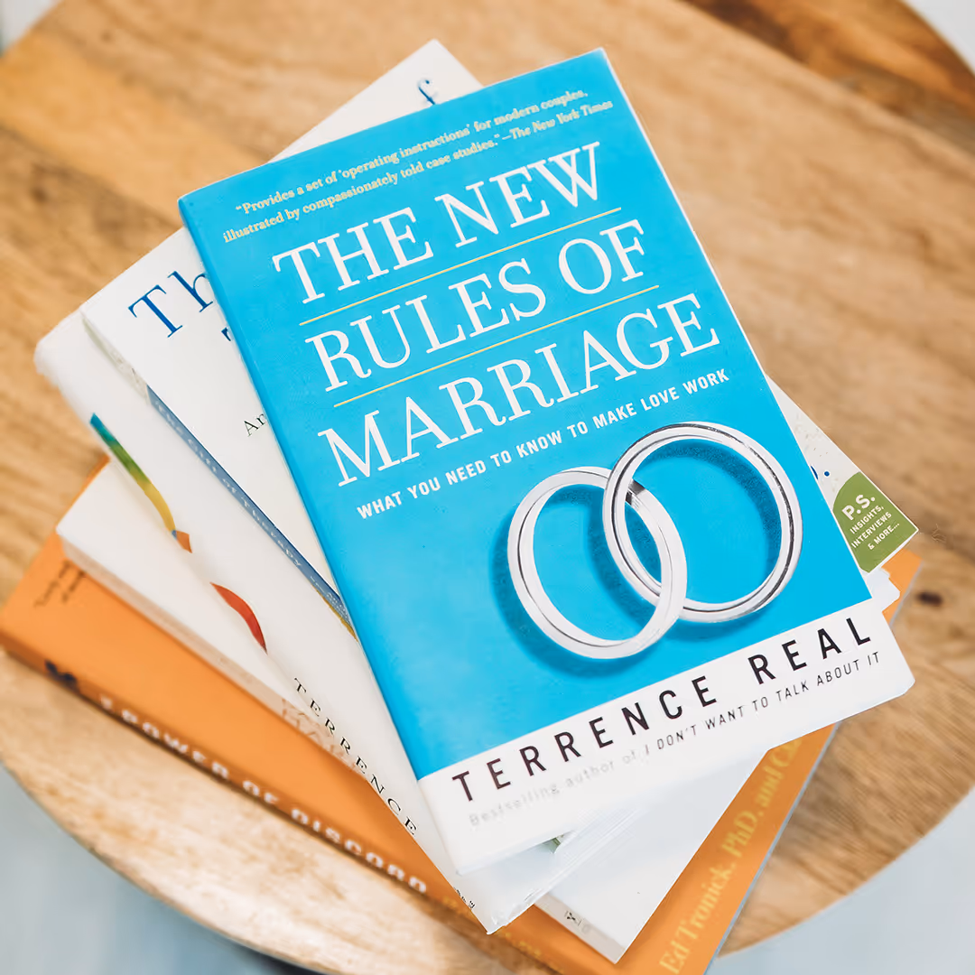 Stack of books on a wooden surface with the top book titled 'The New Rules of Marriage' by Terrence Real featuring two silver wedding rings on the cover.