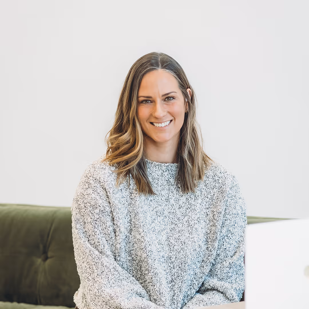 Smiling woman with shoulder-length wavy hair wearing a gray sweater sitting in front of a white background and green couch.