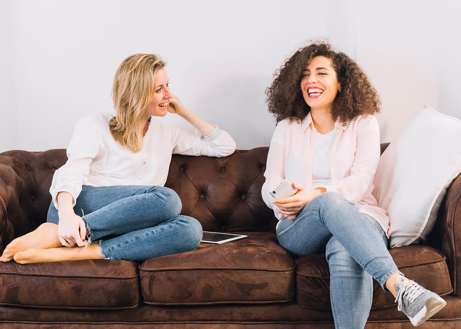 Two women sitting on a brown leather couch, smiling and talking, one holding a smartphone and the other with a tablet beside her.