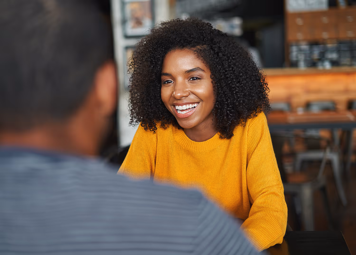 Smiling woman with curly hair wearing a yellow sweater, engaged in conversation with a man in a cafe.