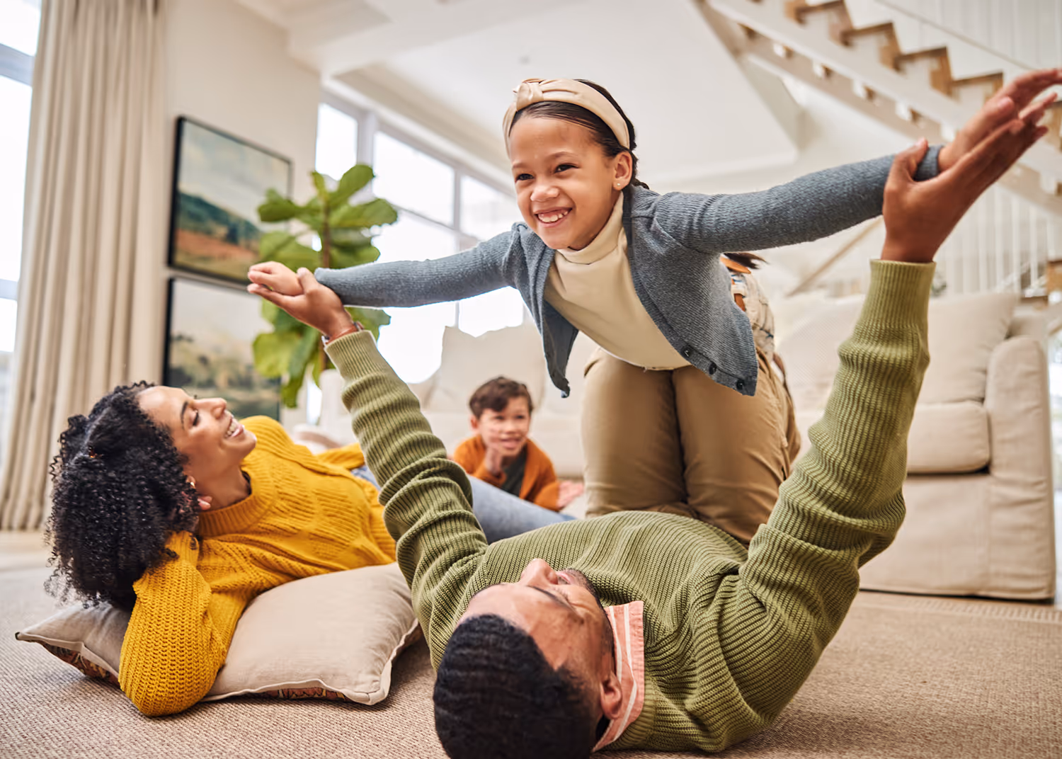 Family playing together indoors, father lying on the floor lifting daughter in the air while mother and son watch with smiles.