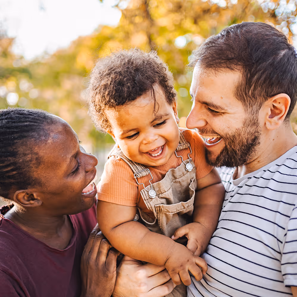 Smiling mixed-race toddler in overalls being held and laughed with by two adults outdoors.