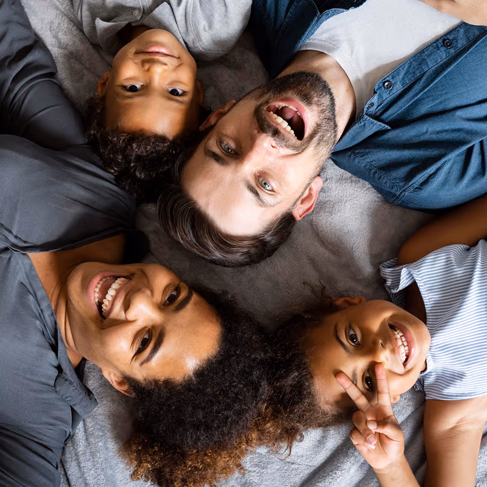 Top view of a smiling diverse family of four lying on a blanket with heads together, one child making a peace sign.