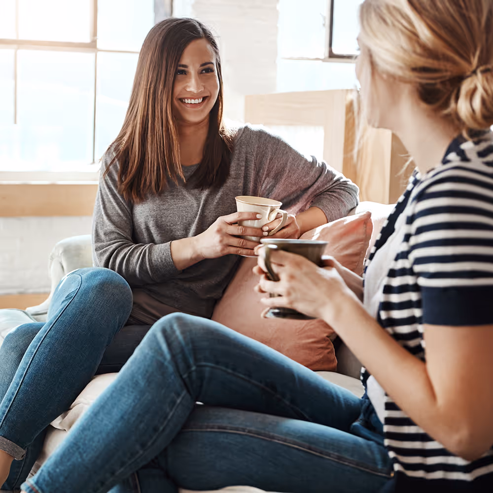 Two women sitting on a couch, smiling and holding coffee mugs while chatting in a bright room.