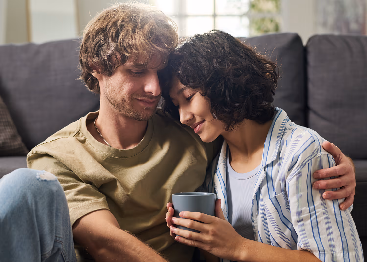 Couple sitting closely on a couch, woman holding a mug and leaning her head on the man's shoulder, both smiling softly.